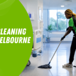 Hospital cleaner mops a bright hallway while wearing a green-and-black uniform; blue 'Patient Rooms' sign visible in the doorway.